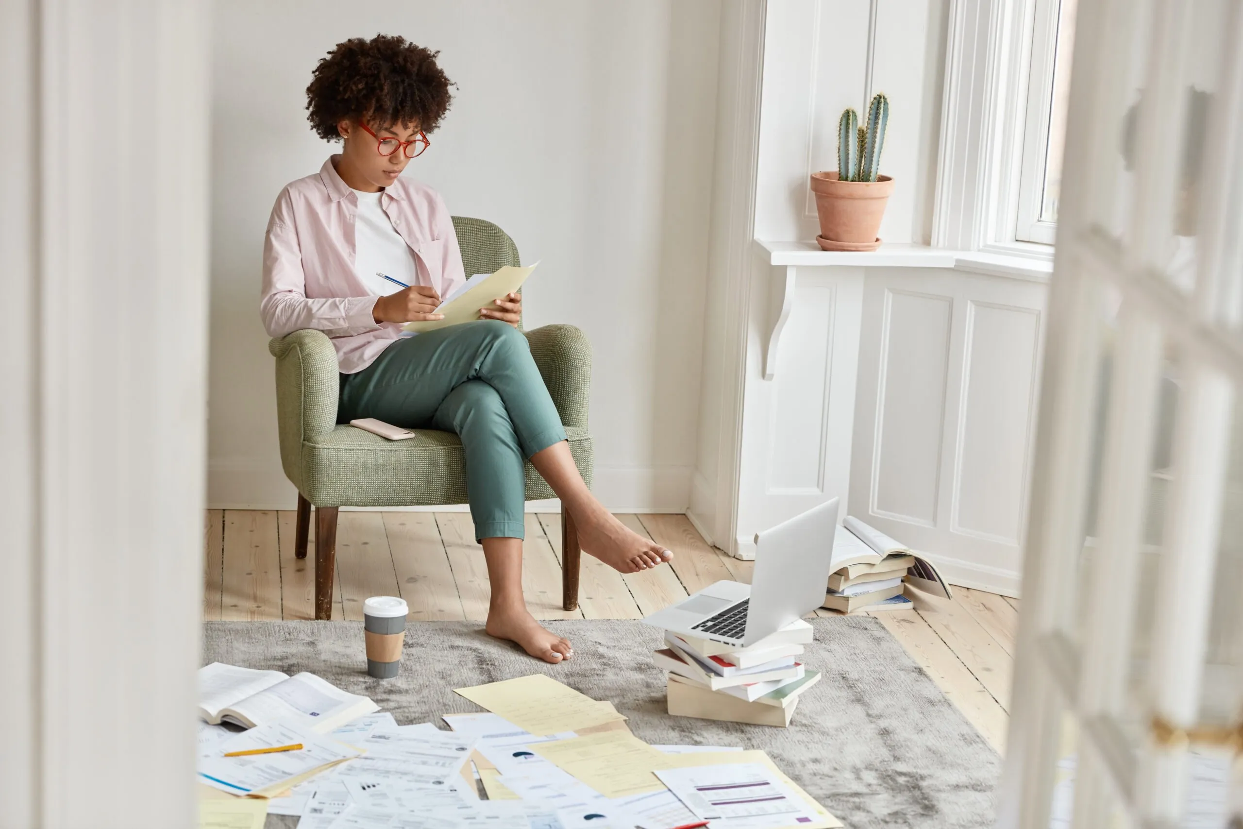 Image of attractive black entrepreneur studies private financing, has disorder in room, involved in work, sits at armchair, manages fund. Busy student prepares for upcoming exams and session